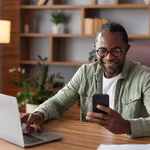 African man involved in gig economy looking at his phone and working on his computer